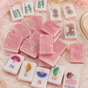 May include: A collection of pink and white Mahjong tiles. The pink tiles are rectangular, while the white tiles feature various colorful designs and symbols. Some tiles display the numbers "1", while others have floral or geometric patterns. The "JOKER" tile is also visible.