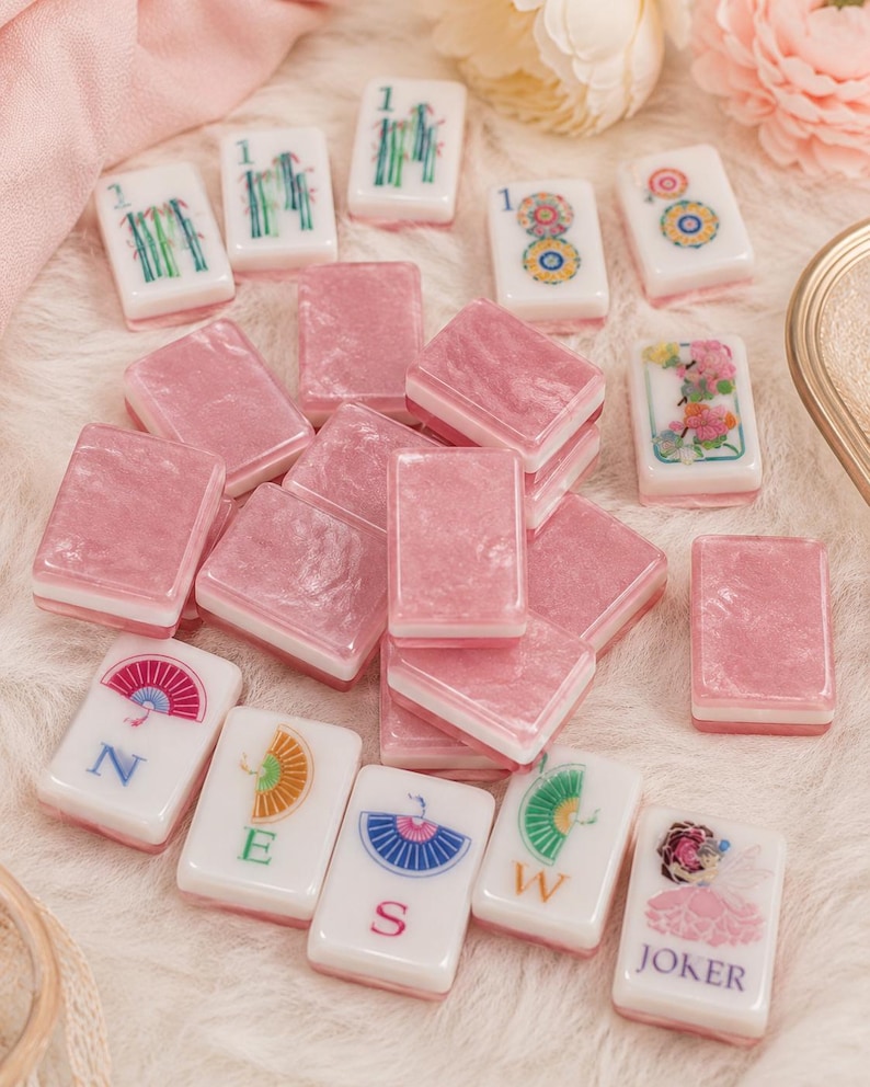 May include: A collection of pink and white Mahjong tiles. The pink tiles are rectangular, while the white tiles feature various colorful designs and symbols. Some tiles display the numbers "1", while others have floral or geometric patterns. The "JOKER" tile is also visible.