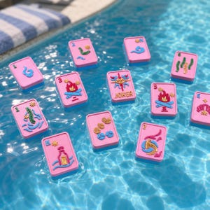 Puede incluir: Cartas de juego rosas y azules flotando en una piscina. Cada carta presenta un dise&ntilde;o &uacute;nico en azul y dorado, con la palabra "JOKER" en una de ellas. Las cartas son rectangulares con esquinas redondeadas.