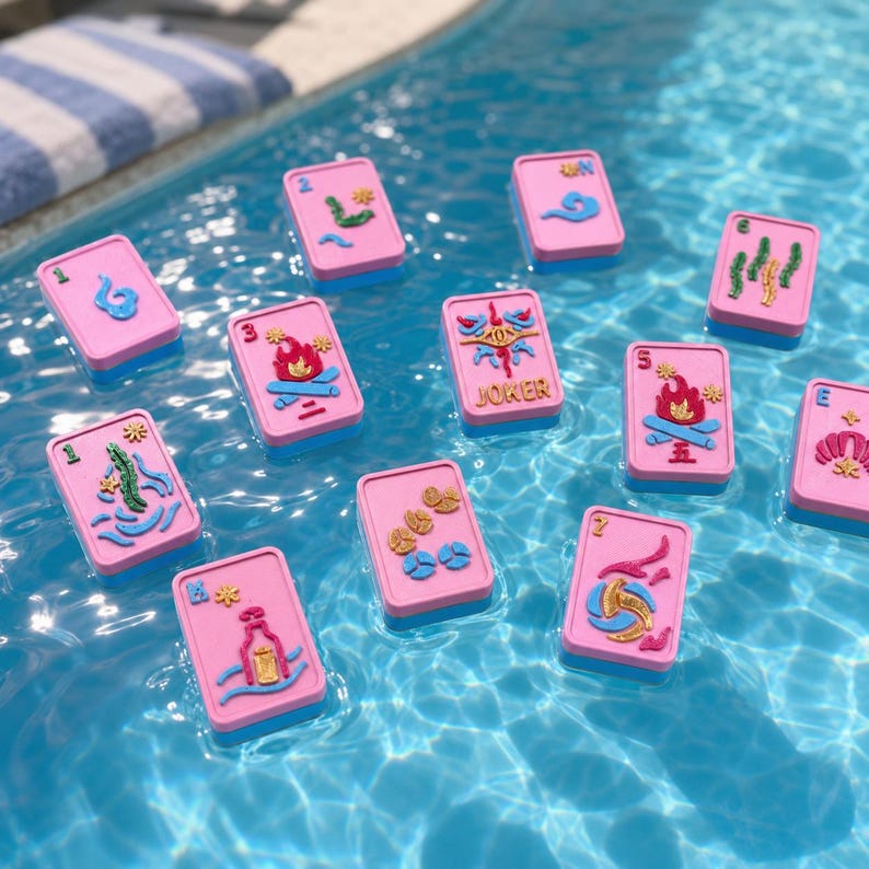 Puede incluir: Cartas de juego rosas y azules flotando en una piscina. Cada carta presenta un dise&ntilde;o &uacute;nico en azul y dorado, con la palabra "JOKER" en una de ellas. Las cartas son rectangulares con esquinas redondeadas.