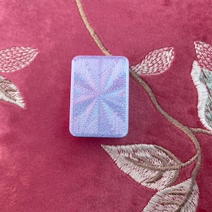 May include: A rectangular, iridescent, light blue object with a swirling pattern. The object is set against a red background with embroidered floral details.
