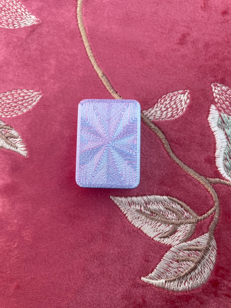 May include: A rectangular, iridescent, light blue object with a swirling pattern. The object is set against a red background with embroidered floral details.