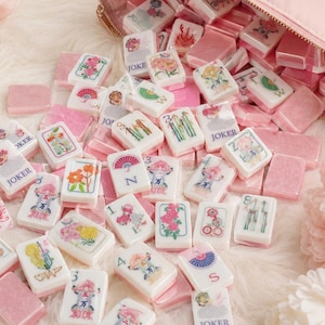May include: A collection of white and pink mahjong tiles with floral and character designs, scattered around a pink zippered pouch. The tiles feature various symbols and the word "JOKER".