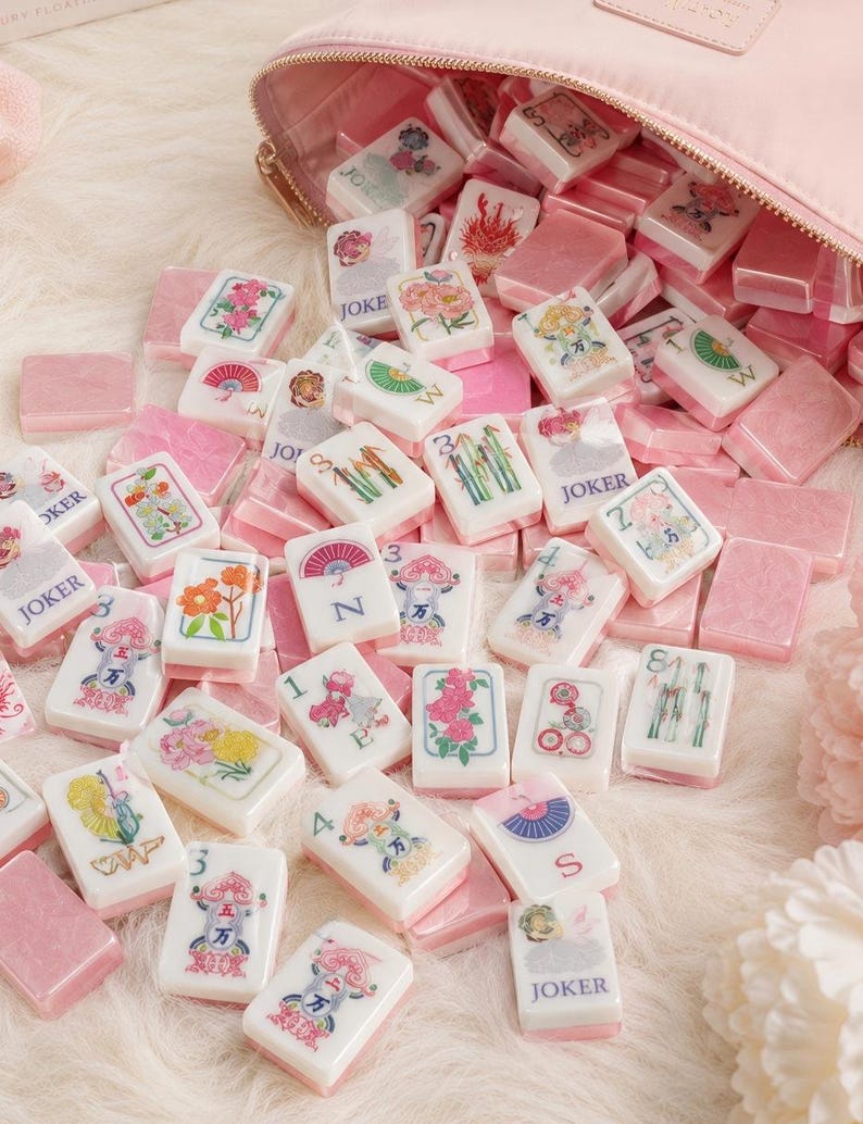 May include: A collection of white and pink mahjong tiles with floral and character designs, scattered around a pink zippered pouch. The tiles feature various symbols and the word "JOKER".