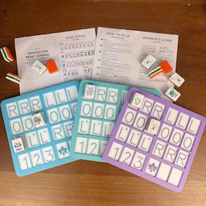 May include: A complete American Mah Jongg set with three colored tile racks, tiles, and instruction sheets. The racks are blue, teal, and purple, each holding white tiles with black lettering and symbols. The set includes orange and green tile holders.