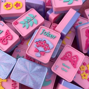 May include: A collection of pink and purple Mahjong tiles with various embossed designs, including flowers, butterflies, and leaves. Some tiles have the word "Joker" on them. The tiles are stacked and scattered, creating a visually appealing arrangement.