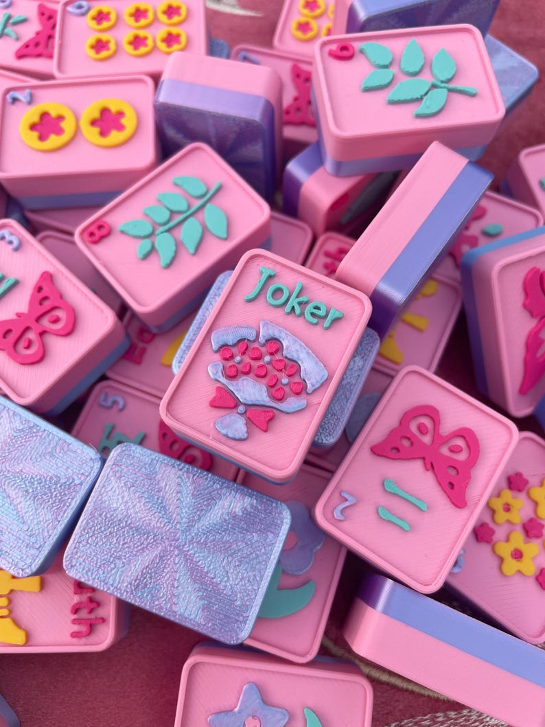 May include: A collection of pink and purple Mahjong tiles with various embossed designs, including flowers, butterflies, and leaves. Some tiles have the word "Joker" on them. The tiles are stacked and scattered, creating a visually appealing arrangement.