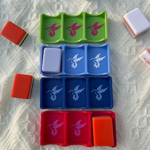 Puede incluir: Una colecci&oacute;n de coloridos soportes para fichas de Mahjong en verde, azul y rojo, cada uno con fichas. Los soportes presentan un dise&ntilde;o de colibr&iacute;. Varias fichas de Mahjong est&aacute;n esparcidas alrededor de los soportes. Las fichas son de color naranja, blanco y pastel.