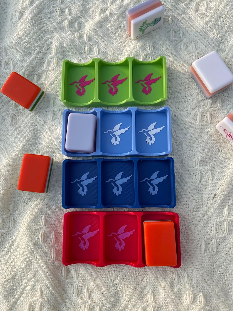 Puede incluir: Una colecci&oacute;n de coloridos soportes para fichas de Mahjong en verde, azul y rojo, cada uno con fichas. Los soportes presentan un dise&ntilde;o de colibr&iacute;. Varias fichas de Mahjong est&aacute;n esparcidas alrededor de los soportes. Las fichas son de color naranja, blanco y pastel.