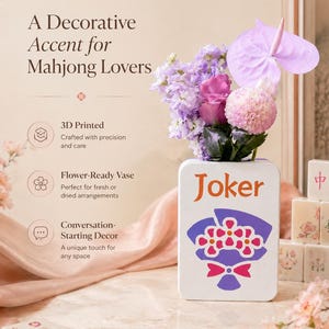 May include: A white, rectangular vase with the word "Joker" in orange, holding a bouquet of purple and pink flowers. The vase has a graphic of flowers and a bow. The text "A Decorative Accent for Mahjong Lovers" is at the top.