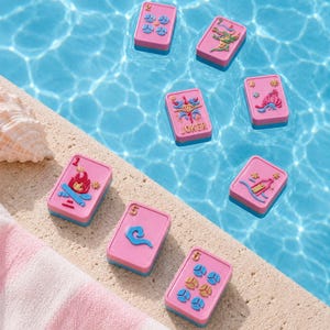 Puede incluir: Cartas de juego rosas y azules flotando en una piscina. Las cartas tienen varios dise&ntilde;os, incluyendo una fogata, un drag&oacute;n y la palabra "JOKER". Una toalla a rayas rosa y blanca y una concha est&aacute;n en el borde de la piscina.