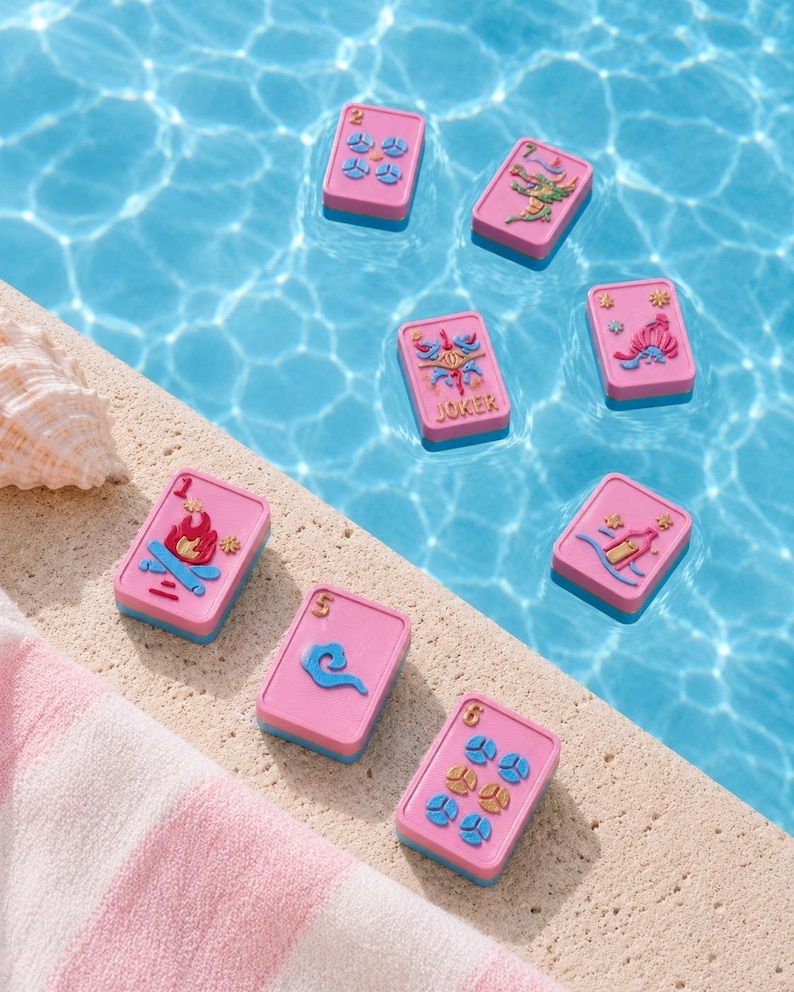 Puede incluir: Cartas de juego rosas y azules flotando en una piscina. Las cartas tienen varios dise&ntilde;os, incluyendo una fogata, un drag&oacute;n y la palabra "JOKER". Una toalla a rayas rosa y blanca y una concha est&aacute;n en el borde de la piscina.