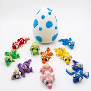 May include: A collection of colorful, handcrafted clay dinosaur figurines and a white egg-shaped container with blue accents. The dinosaurs are in various colors, including red, green, orange, yellow, purple, pink, and blue.
