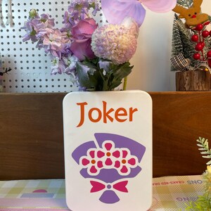 May include: A white rectangular vase with the word "Joker" in orange lettering and a purple and pink floral design. The vase holds a bouquet of purple and pink flowers, including a large anthurium.