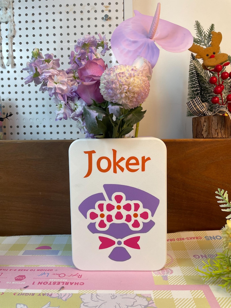 May include: A white rectangular vase with the word "Joker" in orange lettering and a purple and pink floral design. The vase holds a bouquet of purple and pink flowers, including a large anthurium.