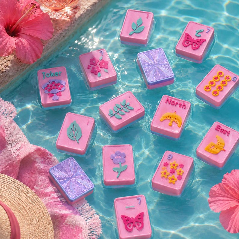 May include: Pink and blue playing cards floating in water. The cards feature various designs, including flowers, leaves, and butterflies. Some cards have the words "Joker", "North", and "East". A straw hat and pink towel are in the foreground.