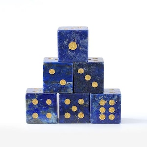 May include: Six blue dice with gold dots are stacked in a pyramid shape. The dice are a deep blue color with gold-colored dots indicating the numbers. The dice are arranged on a white background.