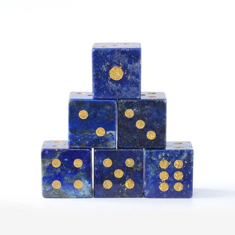 May include: Six blue dice with gold dots are stacked in a pyramid shape. The dice are a deep blue color with gold-colored dots indicating the numbers. The dice are arranged on a white background.