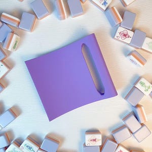 May include: A solid purple square tile rack with a single elongated slot, surrounded by a collection of scattered mahjong tiles in shades of lavender and white, featuring various colorful printed symbols, numbers, and characters.