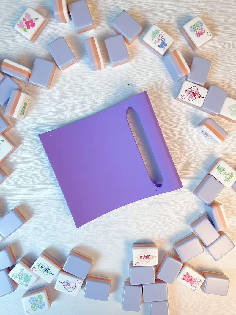 May include: A solid purple square tile rack with a single elongated slot, surrounded by a collection of scattered mahjong tiles in shades of lavender and white, featuring various colorful printed symbols, numbers, and characters.