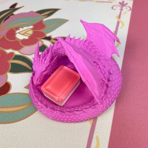 May include: A bright pink 3D-printed dragon figurine curled into a circular shape with textured scales and wings. The dragon is holding a small, rectangular, translucent pink and white block object within its coiled body and wings.