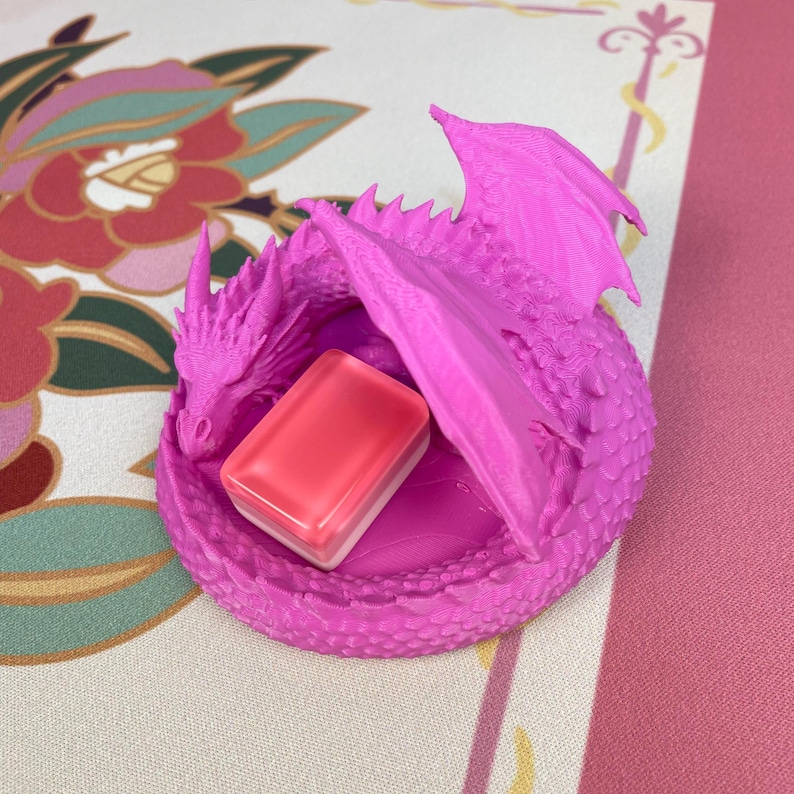 May include: A bright pink 3D-printed dragon figurine curled into a circular shape with textured scales and wings. The dragon is holding a small, rectangular, translucent pink and white block object within its coiled body and wings.