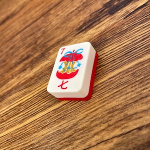 May include: A rectangular mahjong tile with a white face and a red back. The face features the number 7 in red, the Chinese character for seven in red, and a central graphic of a red apple core tied with a blue ribbon and small blue stars.