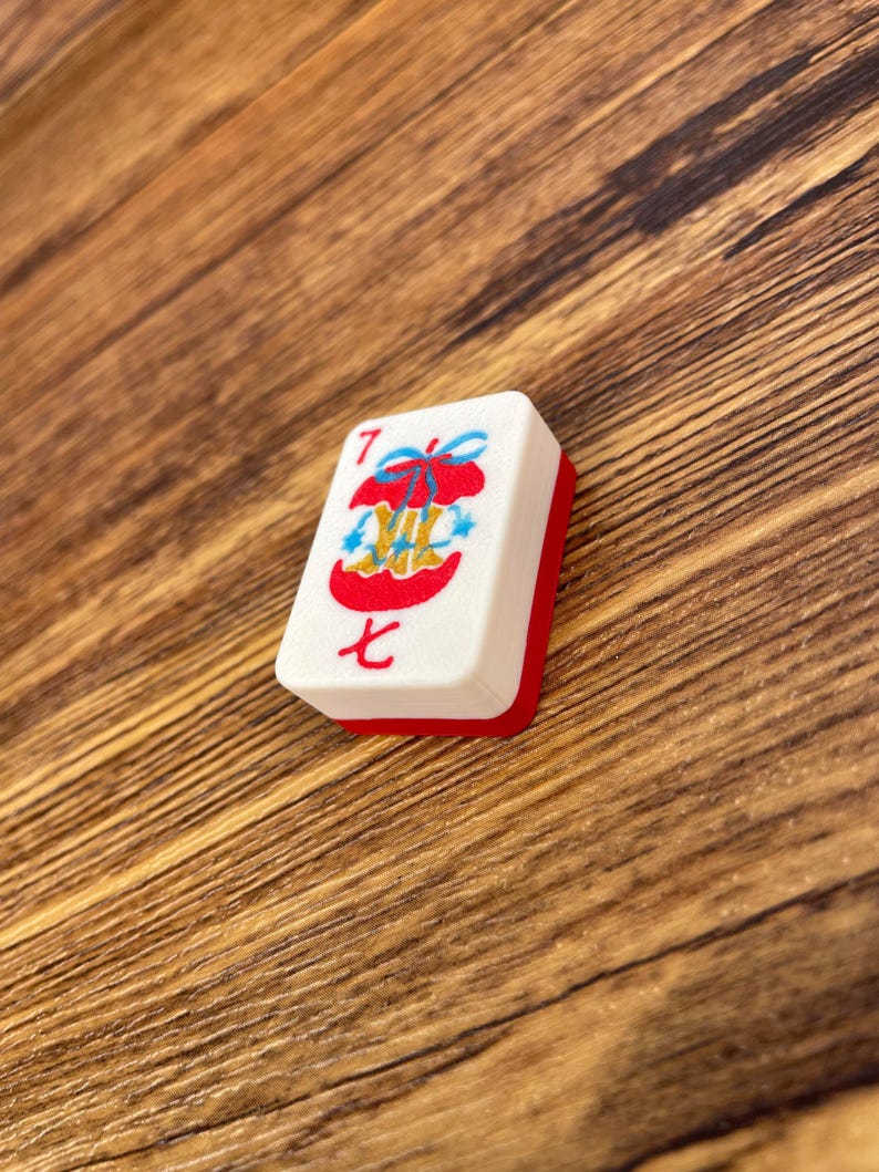 May include: A rectangular mahjong tile with a white face and a red back. The face features the number 7 in red, the Chinese character for seven in red, and a central graphic of a red apple core tied with a blue ribbon and small blue stars.