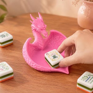 Pu&ograve; includere: Un vassoio rosa brillante a forma di drago con un design alato testurizzato. Il vassoio &egrave; posizionato su una superficie in legno accanto a diverse tessere del mahjong bianche, verdi e arancioni, una delle quali viene posta al centro.