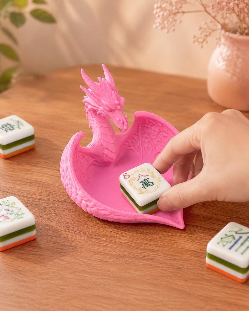 Pu&ograve; includere: Un vassoio rosa brillante a forma di drago con un design alato testurizzato. Il vassoio &egrave; posizionato su una superficie in legno accanto a diverse tessere del mahjong bianche, verdi e arancioni, una delle quali viene posta al centro.
