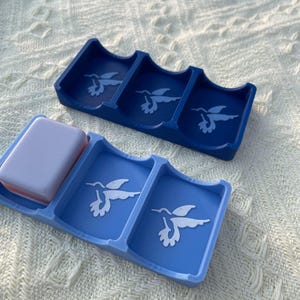 Puede incluir: Dos jaboneras azules con dise&ntilde;os de colibr&iacute;es. Una es azul oscuro, la otra azul claro. Una pastilla de jab&oacute;n descansa en una de las jaboneras. Las jaboneras son de material liso.