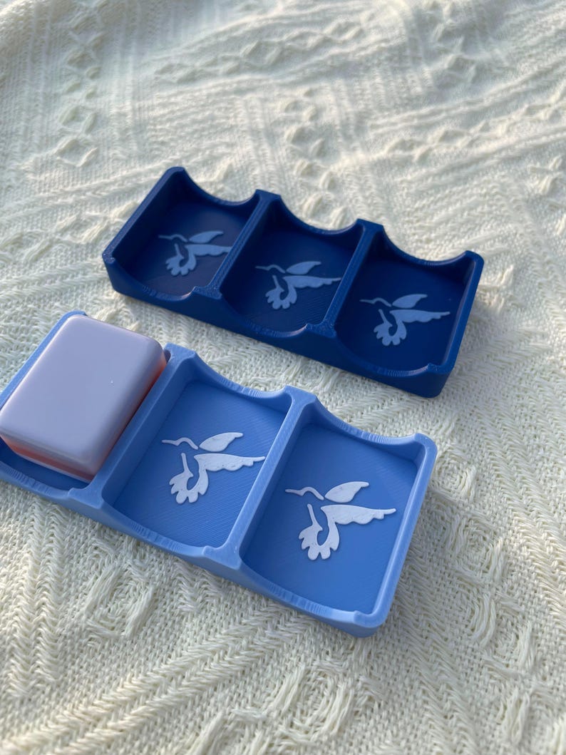 Puede incluir: Dos jaboneras azules con dise&ntilde;os de colibr&iacute;es. Una es azul oscuro, la otra azul claro. Una pastilla de jab&oacute;n descansa en una de las jaboneras. Las jaboneras son de material liso.