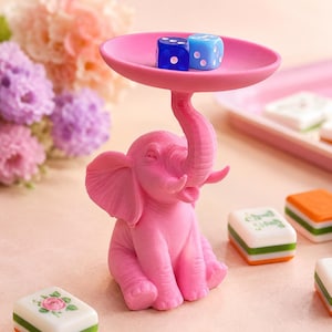 May include: A bright pink elephant figurine with a raised trunk supporting a circular tray. Two translucent dice, one dark blue and one light blue, rest on the tray. The elephant is seated and features textured skin details.