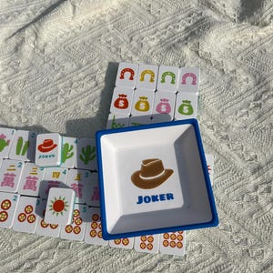 Może przedstawiać: Biała i niebieska kwadratowa taca z brązową grafiką kapelusza kowbojskiego i napisem "JOKER". Taca otoczona jest kafelkami do gry w Mahjonga z r&oacute;żnymi symbolami i numerami. Kafelki są białe z czerwonymi, zielonymi i niebieskimi oznaczeniami.