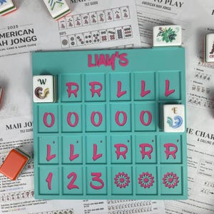 May include: A teal Mahjong tile holder with pink lettering spelling "LIAM'S" and a grid for tiles. The grid contains pink letters, numbers, and flower designs. Several Mahjong tiles and game guides surround the holder.