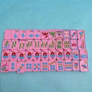 Puede incluir: Un juego de fichas de Mahjong rosas con detalles azules y dorados. Las fichas presentan varios s&iacute;mbolos, n&uacute;meros y caracteres, incluyendo dragones, llamas y la palabra "Joker". Las fichas est&aacute;n dispuestas sobre una superficie azul claro.