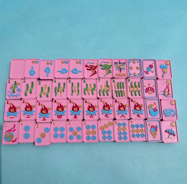 Puede incluir: Un juego de fichas de Mahjong rosas con detalles azules y dorados. Las fichas presentan varios s&iacute;mbolos, n&uacute;meros y caracteres, incluyendo dragones, llamas y la palabra "Joker". Las fichas est&aacute;n dispuestas sobre una superficie azul claro.