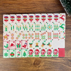 May include: A set of white mahjong tiles featuring Christmas-themed illustrations including reindeer, candles, ornaments, stockings, gifts, and Santa Claus figures. The tiles display numbers, symbols, and the letters S, N, W, and E in various colors.