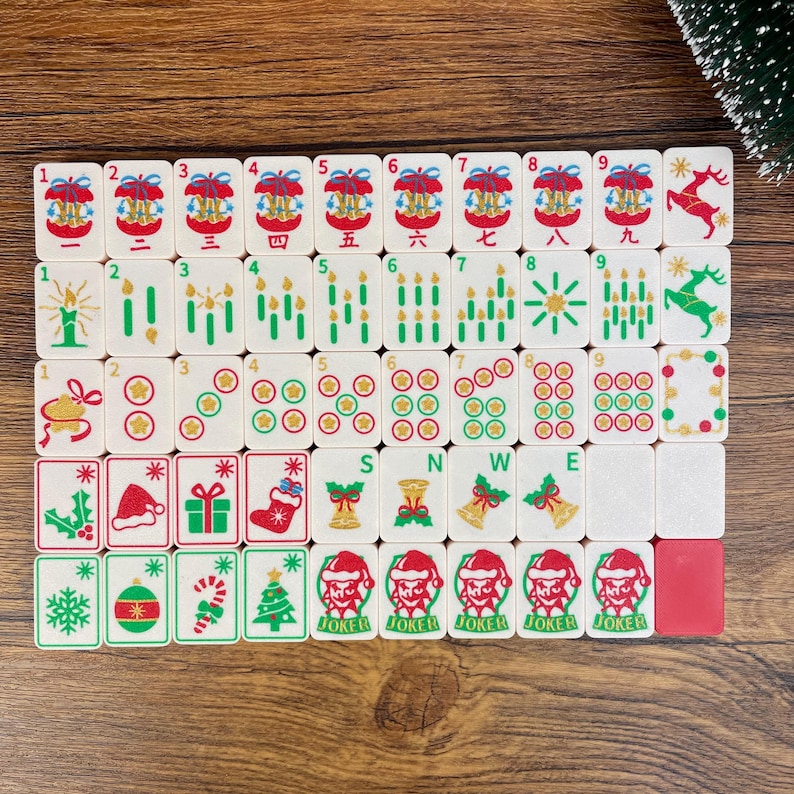 May include: A set of white mahjong tiles featuring Christmas-themed illustrations including reindeer, candles, ornaments, stockings, gifts, and Santa Claus figures. The tiles display numbers, symbols, and the letters S, N, W, and E in various colors.
