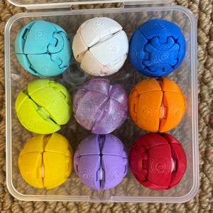 Op de afbeelding: Een doorzichtige plastic doos bevat negen kleurrijke, bolvormige puzzelspeeltjes. De bollen zijn verdeeld in segmenten en zijn er in verschillende kleuren, waaronder turquoise, wit, blauw, groen, paars, oranje, geel en rood. De speeltjes hebben een textuur.