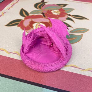 May include: A bright pink 3D printed dragon figurine curled into a circular shape. The dragon features detailed scales, pointed horns, and folded wings, resting its head on its own tail. The base has a textured, rippled surface pattern.