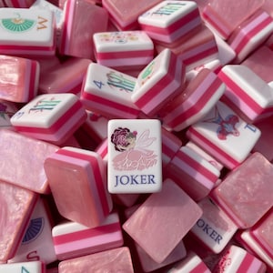May include: A collection of pink and white Mahjong tiles. The tiles feature various symbols, numbers, and the word "JOKER" on one tile. The tiles have pink and white striped edges. The overall aesthetic is a soft, feminine style.