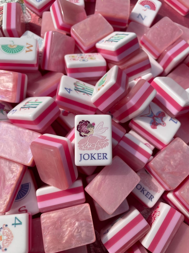 May include: A collection of pink and white Mahjong tiles. The tiles feature various symbols, numbers, and the word "JOKER" on one tile. The tiles have pink and white striped edges. The overall aesthetic is a soft, feminine style.