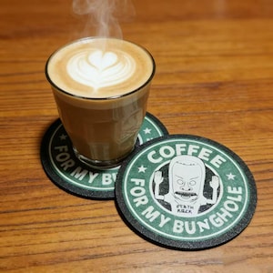 May include: A glass of latte with a heart-shaped design in the foam sits atop two green and black coasters. The coasters feature a cartoon character and the text "COFFEE FOR MY BUNGHOLE."