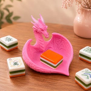 Pu&ograve; includere: Un vassoio portagioie rosa brillante a forma di drago con ali testurizzate e una conca centrale, che contiene una tessera del mahjong arancione, bianca e verde. Quattro tessere del mahjong con caratteri verdi e neri circondano il drago.