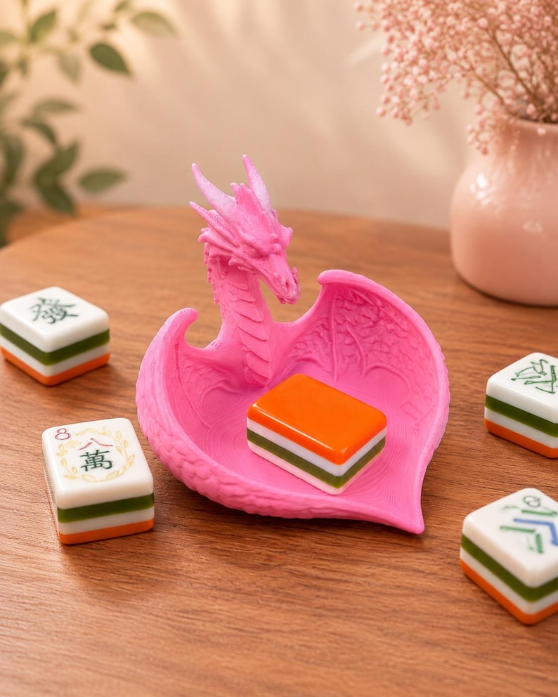 Pu&ograve; includere: Un vassoio portagioie rosa brillante a forma di drago con ali testurizzate e una conca centrale, che contiene una tessera del mahjong arancione, bianca e verde. Quattro tessere del mahjong con caratteri verdi e neri circondano il drago.