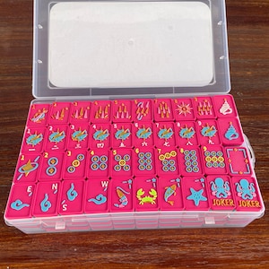 Puede incluir: Un juego de fichas de mahjong de color rosa brillante dispuestas en un estuche de pl&aacute;stico transparente. Las fichas presentan dise&ntilde;os coloridos en relieve que incluyen n&uacute;meros, caracteres chinos, criaturas marinas y la palabra JOKER.