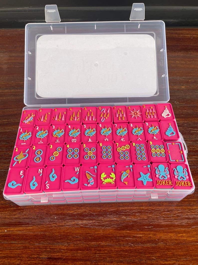 Puede incluir: Un juego de fichas de mahjong de color rosa brillante dispuestas en un estuche de pl&aacute;stico transparente. Las fichas presentan dise&ntilde;os coloridos en relieve que incluyen n&uacute;meros, caracteres chinos, criaturas marinas y la palabra JOKER.