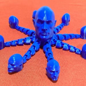 May include: A blue, 3D-printed octopus-like sculpture. The central head is surrounded by eight articulated tentacles, each ending in a human head. The sculpture is a vibrant blue color and sits on an orange surface.
