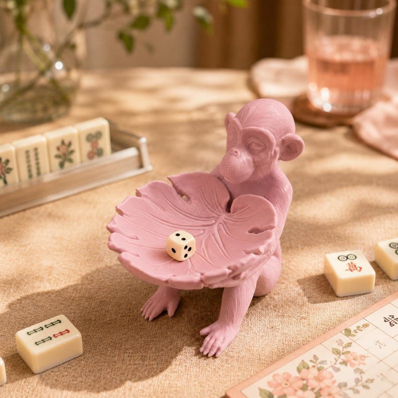 May include: A matte pink monkey figurine holding a large, textured leaf-shaped bowl. A single white six-sided die with black pips rests inside the leaf bowl. The monkey is surrounded by several cream-colored mahjong tiles on a tan surface.
